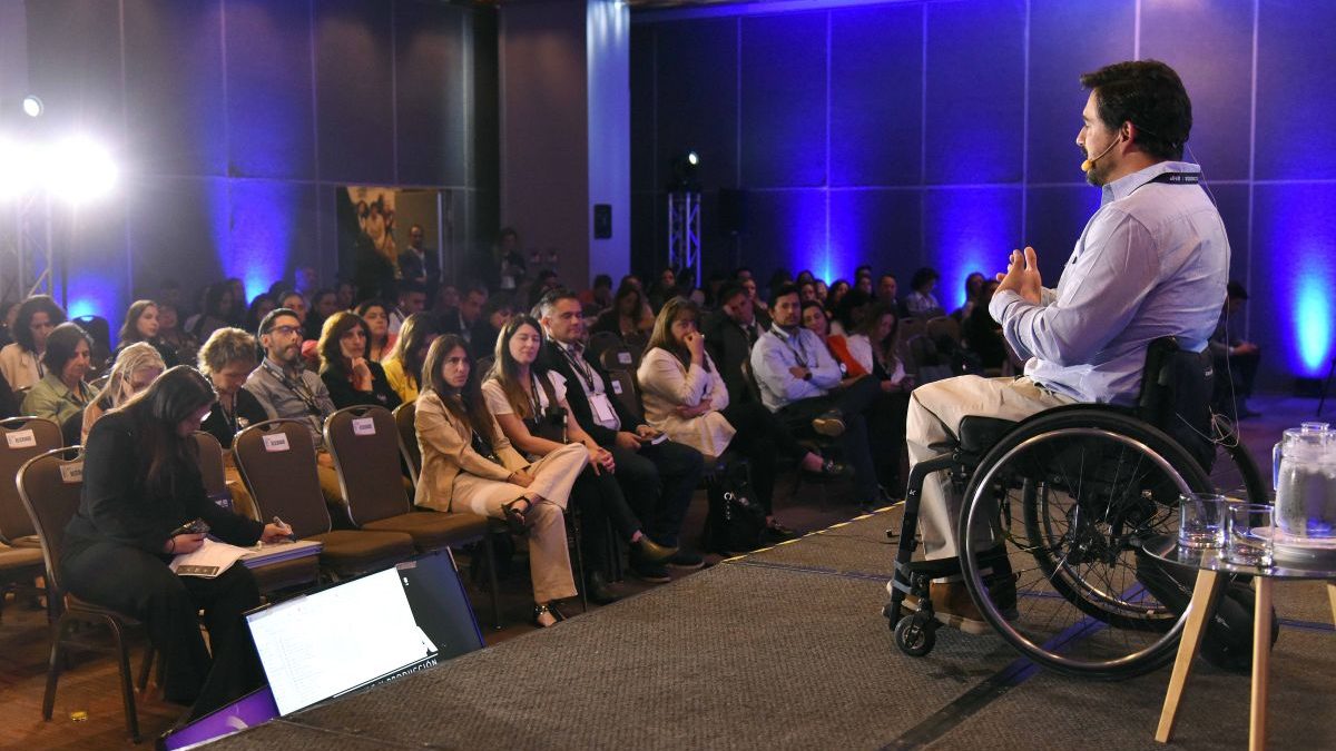 Hombre en silla de ruedas, usa una casa azul, un pantalón beige y está sobre un escenario dirigiéndose hacia una audiencia sentada en sillas en el congreso Summit DEI.