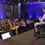 Hombre en silla de ruedas, usa una casa azul, un pantalón beige y está sobre un escenario dirigiéndose hacia una audiencia sentada en sillas en el congreso Summit DEI.