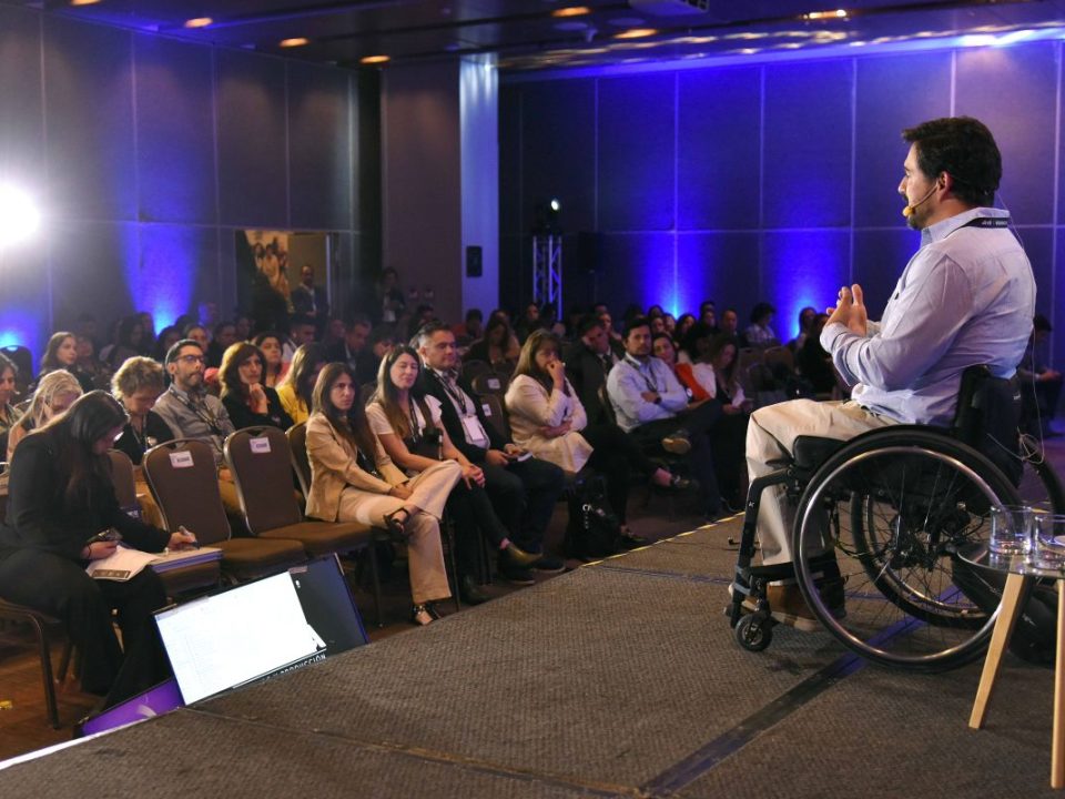 Hombre en silla de ruedas, usa una casa azul, un pantalón beige y está sobre un escenario dirigiéndose hacia una audiencia sentada en sillas en el congreso Summit DEI.